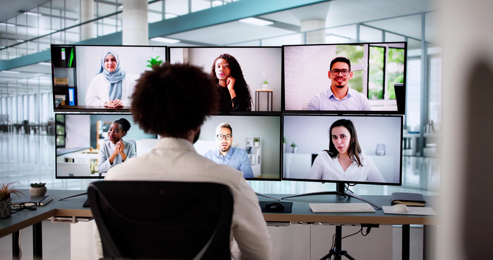 A man sits in a chair, back to the camera. Before him are faces of diverse people on a monitor.