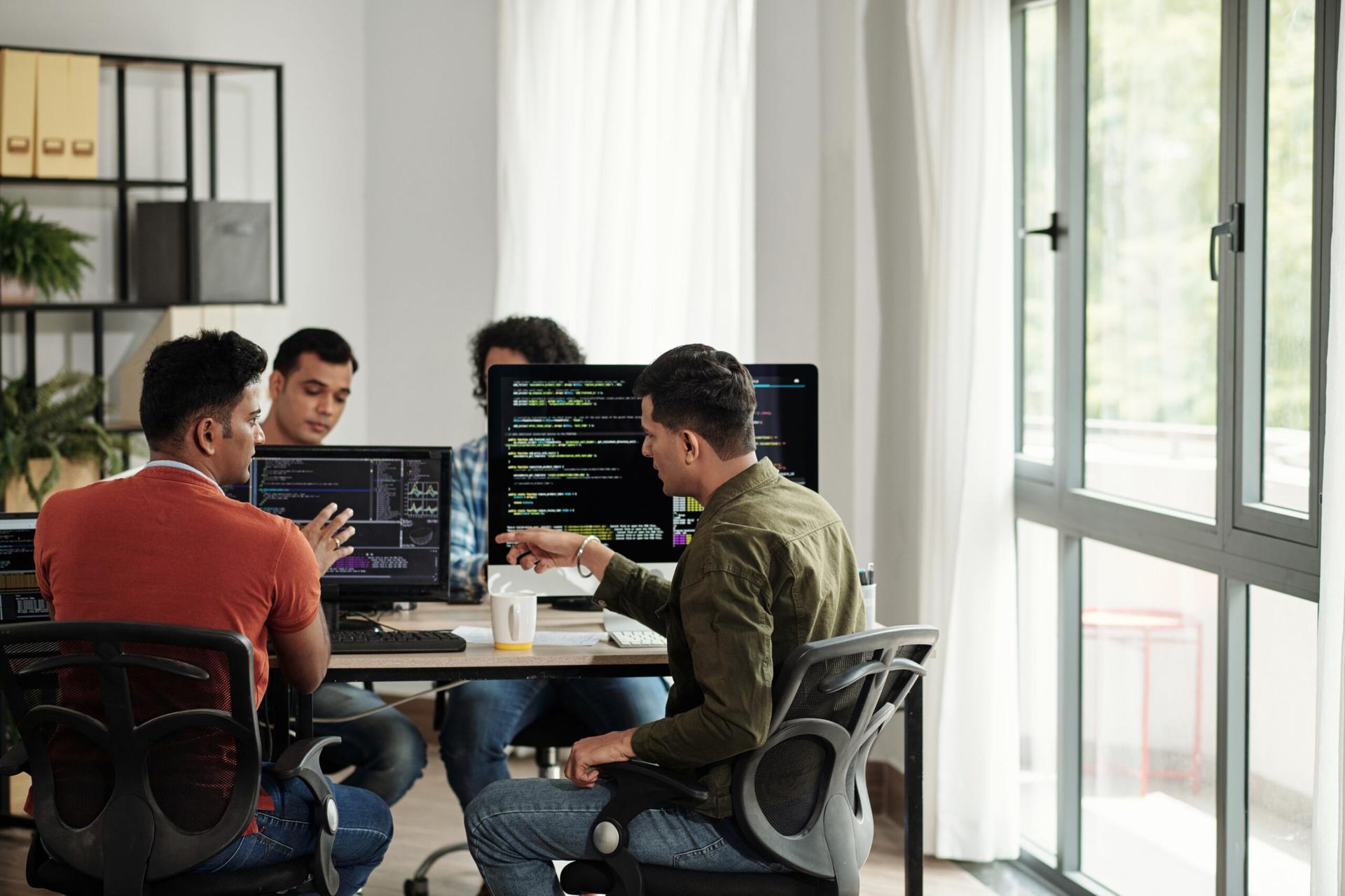 Group of 4 developers sitting at their desktop setups talking